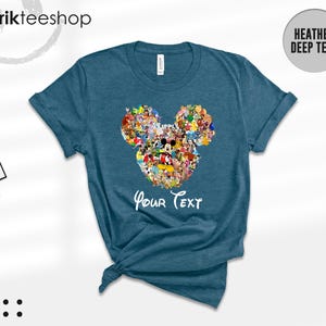 Pu&ograve; includere: T-shirt color teal melange con un design a forma di testa di Topolino composto da vari personaggi dei cartoni animati. Le parole "Your Text" sono scritte in bianco sotto il disegno. La maglietta &egrave; piegata e annodata in basso.