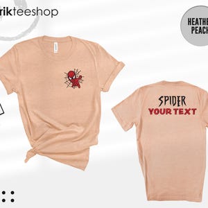 May include: A heather peach short-sleeve t-shirt featuring a small red and black spider-themed character graphic on the front left chest. The back of the shirt displays the text SPIDER and YOUR TEXT in a bold, dark, stylized font.