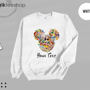 Pu&ograve; includere: Felpa bianca con un colorato design a forma di testa di Topolino composto da vari personaggi dei cartoni animati. Le parole "Your Text" sono scritte in un carattere script sotto il design. La felpa ha un girocollo.