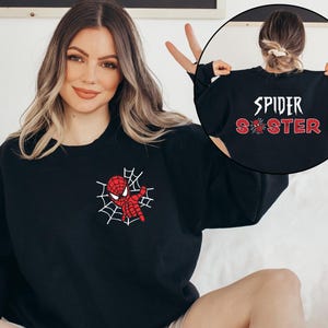 May include: A black crewneck sweatshirt featuring a small red and white spider web graphic on the left chest. The back of the sweatshirt displays the white text SPIDER above the red glittery text SISTER, with a small spider icon replacing the letter I.