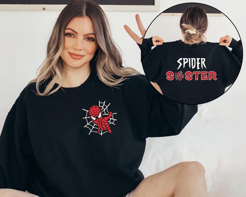 May include: A black crewneck sweatshirt featuring a small red and white spider web graphic on the left chest. The back of the sweatshirt displays the white text SPIDER above the red glittery text SISTER, with a small spider icon replacing the letter I.