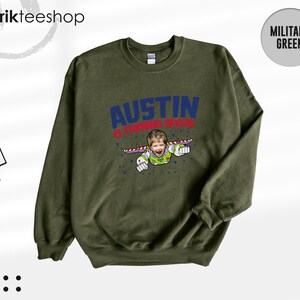 K&ouml;nnte beinhalten: Ein milit&auml;rgr&uuml;ner Rundhals-Pullover mit einer Grafik, die das Gesicht eines Kindes auf dem K&ouml;rper einer Weltraumfigur zeigt. Der Text AUSTIN IS TURNING SEVEN ist in blauen und roten Buchstaben gedruckt, umgeben von kleinen blauen Sternen.