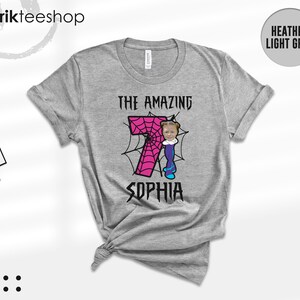 May include: A heather light grey t-shirt featuring a graphic with the text THE AMAZING and SOPHIA in a jagged black font. A large pink number 7 with a black spiderweb pattern is centered, accompanied by a cartoon figure with a photo-realistic face.