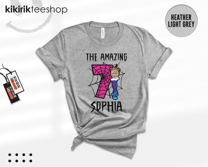 May include: A heather light grey t-shirt featuring a graphic with the text THE AMAZING and SOPHIA in a jagged black font. A large pink number 7 with a black spiderweb pattern is centered, accompanied by a cartoon figure with a photo-realistic face.