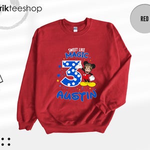 May include: A red crewneck sweatshirt featuring a graphic print with the text SWEET LIKE MAGIC, the number 3 in blue with white polka dots, and the name AUSTIN in blue. A cartoon character with a child's face is positioned next to the number 3.