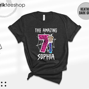 May include: A heather dark grey t-shirt featuring a graphic with a large pink number seven filled with a spiderweb pattern. The design includes a cartoon body with a child's face, white spiderwebs, and the text THE AMAZING SOPHIA.