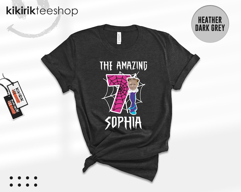 May include: A heather dark grey t-shirt featuring a graphic with a large pink number seven filled with a spiderweb pattern. The design includes a cartoon body with a child's face, white spiderwebs, and the text THE AMAZING SOPHIA.