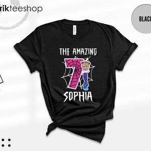 May include: A black short-sleeve t-shirt featuring a graphic with the text THE AMAZING SOPHIA. The design includes a large pink number 7 with a spiderweb pattern, a cartoon character body, and a photograph of a child's face on the character.