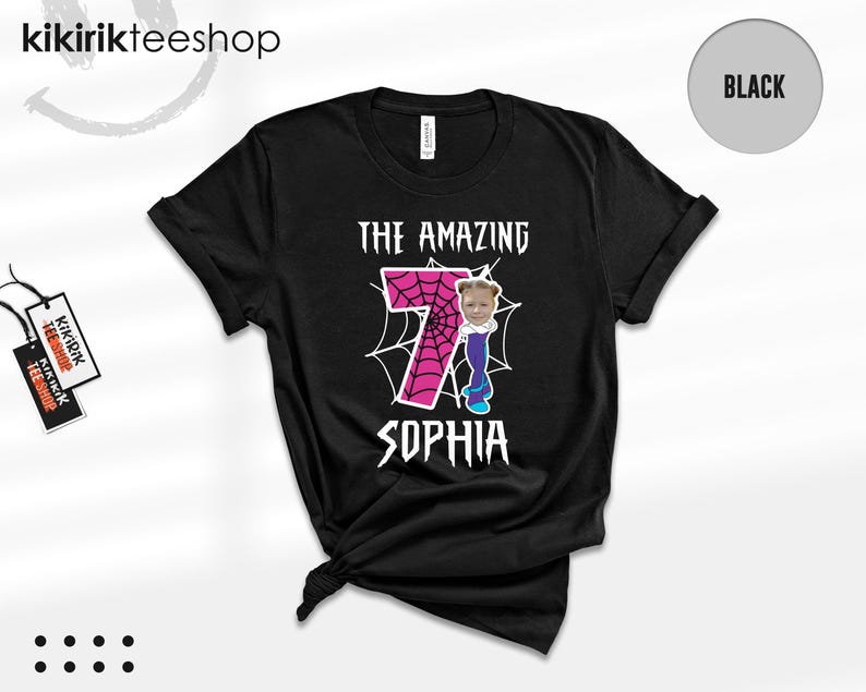May include: A black short-sleeve t-shirt featuring a graphic with the text THE AMAZING SOPHIA. The design includes a large pink number 7 with a spiderweb pattern, a cartoon character body, and a photograph of a child's face on the character.