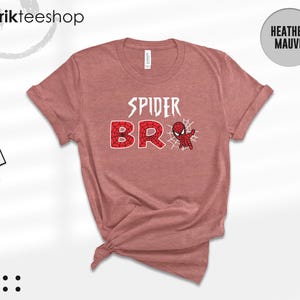 May include: A heather mauve short-sleeve t-shirt featuring the white text SPIDER and the red web-patterned text BRO, accompanied by a small red and blue spider-themed character graphic and a white spider web design on the front center.