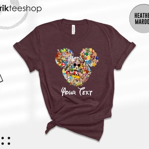 Pu&ograve; includere: T-shirt bordeaux con un design a forma di testa di Topolino riempito di vari personaggi dei cartoni animati. Le parole "Your Text" sono scritte in bianco sotto il disegno. La maglietta ha le maniche arrotolate.