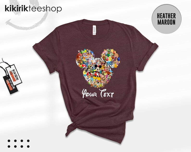 Pu&ograve; includere: T-shirt bordeaux con un design a forma di testa di Topolino riempito di vari personaggi dei cartoni animati. Le parole "Your Text" sono scritte in bianco sotto il disegno. La maglietta ha le maniche arrotolate.