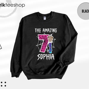 May include: A black long-sleeve sweatshirt featuring a graphic print with the text THE AMAZING SOPHIA. The design includes a large pink number seven with a black spiderweb pattern, and a small cartoon figure with a photo of a child's face.