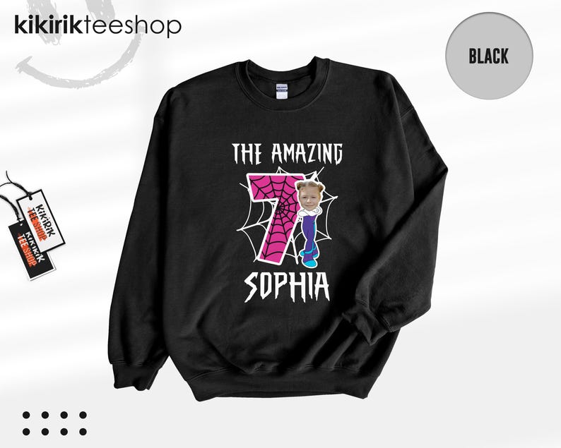 May include: A black long-sleeve sweatshirt featuring a graphic print with the text THE AMAZING SOPHIA. The design includes a large pink number seven with a black spiderweb pattern, and a small cartoon figure with a photo of a child's face.