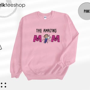 May include: A light pink crewneck sweatshirt featuring the text THE AMAZING MOM in black lettering. The letter O is replaced by a spiderweb graphic with a small cartoon character in a blue suit and white gloves positioned in the center.