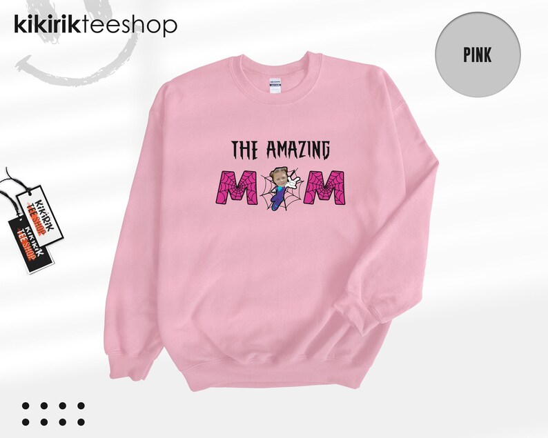 May include: A light pink crewneck sweatshirt featuring the text THE AMAZING MOM in black lettering. The letter O is replaced by a spiderweb graphic with a small cartoon character in a blue suit and white gloves positioned in the center.