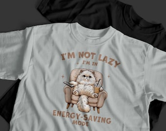 May include: White t-shirt with the text "I'M NOT LAZY I'M IN ENERGY-SAVING MODE." The graphic shows a fluffy cat in a chair holding a remote and a drink. A black t-shirt is visible in the background.