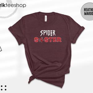 Puede incluir: Camiseta color granate jaspeado con las palabras "SPIDER SISTER" en letras blancas y rojas. La palabra "SPIDER" est&aacute; en negrita, y "SISTER" en un estilo desgastado. Un gr&aacute;fico de ara&ntilde;a est&aacute; entre las palabras, sobre un fondo granate.