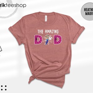 May include: A heather mauve short-sleeve t-shirt featuring the text THE AMAZING DAD in white. The letter A is replaced by a graphic of a child in a blue superhero costume, and the letters D are filled with a pink spiderweb pattern.