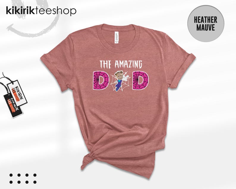 May include: A heather mauve short-sleeve t-shirt featuring the text THE AMAZING DAD in white. The letter A is replaced by a graphic of a child in a blue superhero costume, and the letters D are filled with a pink spiderweb pattern.