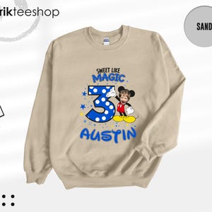 May include: A sand-colored crewneck sweatshirt featuring a graphic print with the text SWEET LIKE MAGIC, the number 3 in blue with white polka dots, and the name AUSTIN. A cartoon character with a child's face is positioned next to the number 3.