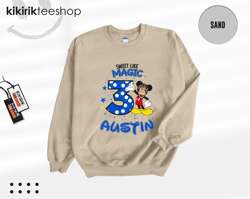 May include: A sand-colored crewneck sweatshirt featuring a graphic print with the text SWEET LIKE MAGIC, the number 3 in blue with white polka dots, and the name AUSTIN. A cartoon character with a child's face is positioned next to the number 3.