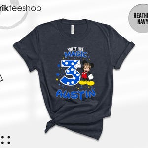 May include: A heather navy short-sleeve t-shirt featuring a graphic with the text "SWEET LIKE MAGIC" above a large blue polka-dot number "3" and the name "AUSTIN" below. A cartoon character with a child's face is positioned next to the number.