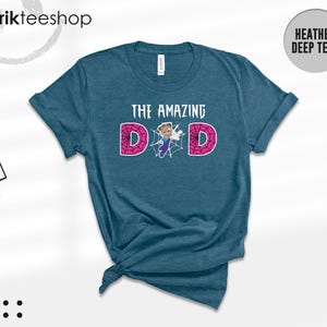 May include: A heather deep teal short-sleeve t-shirt featuring the text THE AMAZING DAD in white. The letter A is replaced by a cartoon character in a blue and purple suit, and the two D letters are filled with a pink spider web pattern.