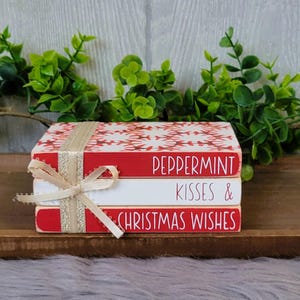 May include: A stack of three red and white decorative books with a gold ribbon. The top book says "Peppermint", the middle book says "Kisses &", and the bottom book says "Christmas Wishes".