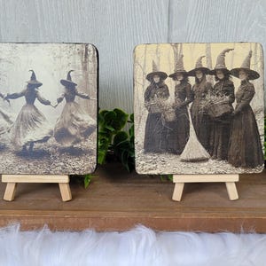Puede incluir: Dos placas cuadradas de madera con im&aacute;genes en sepia de brujas. Una muestra a tres brujas bailando en un bosque, la otra representa a un grupo de brujas con una escoba. Cada placa se exhibe en un peque&ntilde;o caballete de madera.
