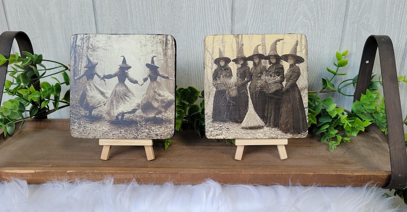 Puede incluir: Dos placas cuadradas de madera con im&aacute;genes en sepia de brujas. Una muestra a tres brujas bailando en un bosque, la otra representa a un grupo de brujas con una escoba. Cada placa se exhibe en un peque&ntilde;o caballete de madera.