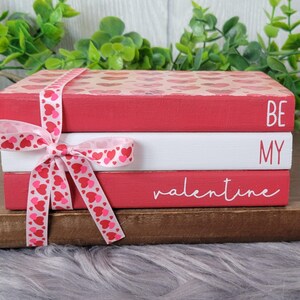 May include: Three stacked red and white books with a pink and white ribbon tied around them. The top book says "Be My Valentine" in white lettering.