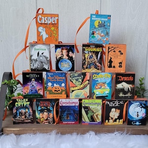 May include: A collection of miniature Halloween-themed book ornaments. Titles include "Casper", "The Haunted House", "The Goonies", "Frankenstein", "Scooby Doo", and "Dracula". Each book has an orange ribbon for hanging and is displayed on a wooden shelf.