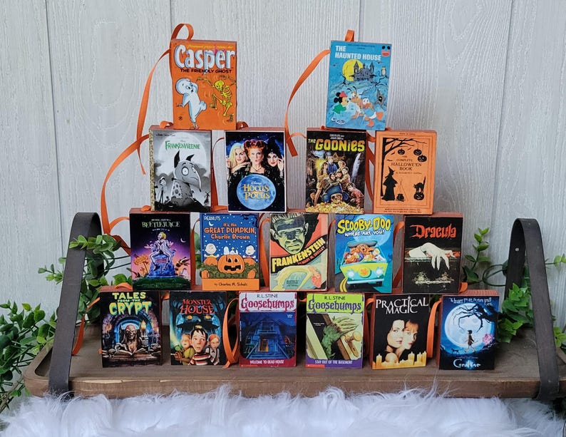 May include: A collection of miniature Halloween-themed book ornaments. Titles include "Casper", "The Haunted House", "The Goonies", "Frankenstein", "Scooby Doo", and "Dracula". Each book has an orange ribbon for hanging and is displayed on a wooden shelf.