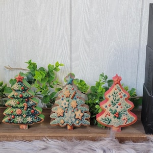 May include: Wooden Christmas tree ornaments with a green, red, and blue color scheme. The ornaments are decorated with stars, candy canes, and holly. The ornaments are displayed on a wooden tray with green foliage. The word "JOY" is spelled out in black and white on wooden blocks.