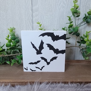 May include: A white square wooden block featuring a black bat silhouette design. The bats are in various sizes and positions, creating a dynamic, Halloween-themed decoration. The block is displayed on a wooden shelf with greenery.