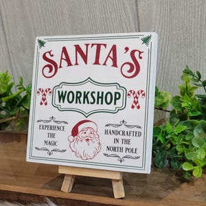 May include: White wooden sign with red and green text that reads "Santa's Workshop". The sign also features a cartoon image of Santa Claus, candy canes, and the text "Experience the Magic" and "Handcrafted in the North Pole".
