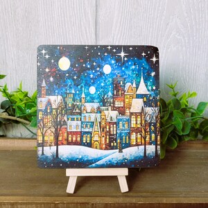 May include: A painting of a winter village scene with colorful houses, snow-covered ground, and a starry night sky. The painting is on a small easel.