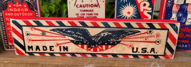 May include: Red, white, and blue wooden sign with a bald eagle and fireworks. The sign reads "Made in U.S.A."