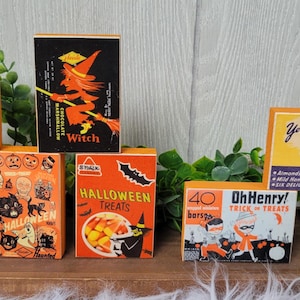 May include: A collection of vintage-style Halloween-themed candy boxes. The boxes feature retro graphics and text, including "Baby Ruth," "Witch," "Halloween Treats," "Oh Henry!," and "Dots." The color palette is dominated by orange, black, and yellow.