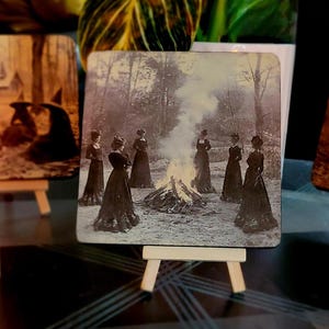 Puede incluir: Posavasos cuadrado con una imagen vintage en blanco y negro de mujeres con vestidos largos reunidas alrededor de una hoguera en un bosque. El posavasos se exhibe en un peque&ntilde;o caballete de madera, con otros posavasos y velas en el fondo.