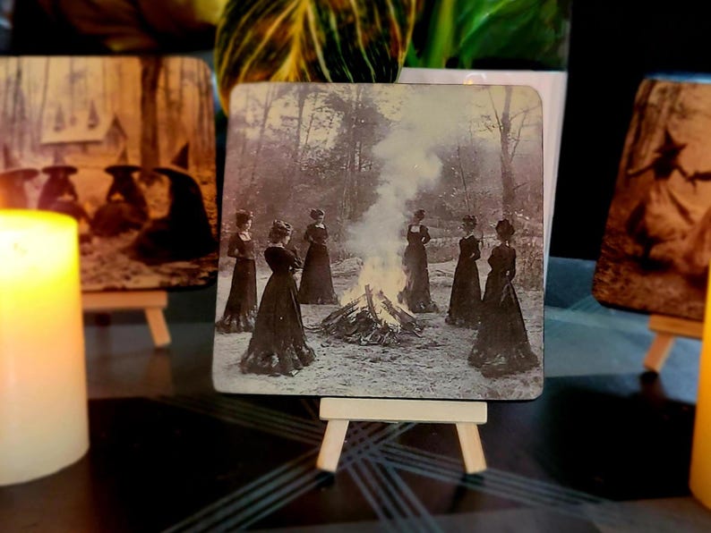 Puede incluir: Posavasos cuadrado con una imagen vintage en blanco y negro de mujeres con vestidos largos reunidas alrededor de una hoguera en un bosque. El posavasos se exhibe en un peque&ntilde;o caballete de madera, con otros posavasos y velas en el fondo.