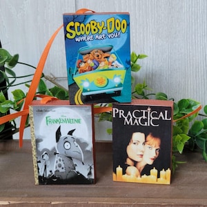 May include: Three miniature book ornaments. The top ornament features the Scooby-Doo cartoon characters in a van. The bottom left ornament is a Frankenweenie book cover. The bottom right ornament is a Practical Magic book cover.