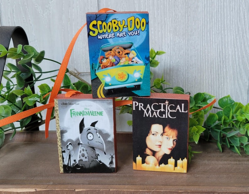 May include: Three miniature book ornaments. The top ornament features the Scooby-Doo cartoon characters in a van. The bottom left ornament is a Frankenweenie book cover. The bottom right ornament is a Practical Magic book cover.