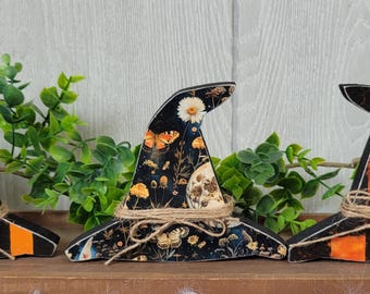 May include: Three wooden witch hats with different designs. One has orange and black stripes, another features a floral and moon pattern, and the third has an orange and black checkerboard pattern. Each hat is tied with twine.