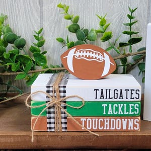 May include: Three wooden signs with football-themed words and graphics. The first sign says "TOUCHDOWN KICKOFF INTERCEPTION FIELD GOAL DEFENSE QUARTERBACK END ZONE BLITZ FIRST & TEN". The second sign has a brown football and the words "TAILGATES TACKLES TOUCHDOWNS". The third sign has a black and white silhouette of a football player and the word "FOOTBALL".