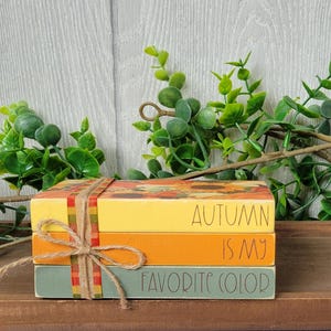 May include: A stack of three decorative wooden blocks tied with twine. The top block is yellow with a sunflower design and the words "AUTUMN" printed on it. The middle block is orange with "IS MY" and the bottom block is green with "FAVORITE COLOR".