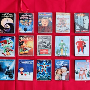 May include: A collection of miniature Christmas movie VHS tape ornaments, including "The Nightmare Before Christmas," "Home Alone," and "Elf." Each ornament features colorful artwork and a red ribbon for hanging. The ornaments are arranged on a red surface.