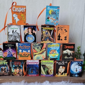 May include: A collection of miniature Halloween-themed book ornaments, including titles like "Casper," "The Haunted House," and "Goosebumps." Each book has a colorful cover and an orange ribbon for hanging. The books are arranged on a wooden tray.