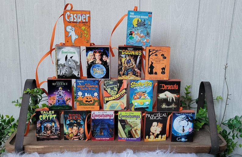 May include: A collection of miniature Halloween-themed book ornaments, including titles like "Casper," "The Haunted House," and "Goosebumps." Each book has a colorful cover and an orange ribbon for hanging. The books are arranged on a wooden tray.